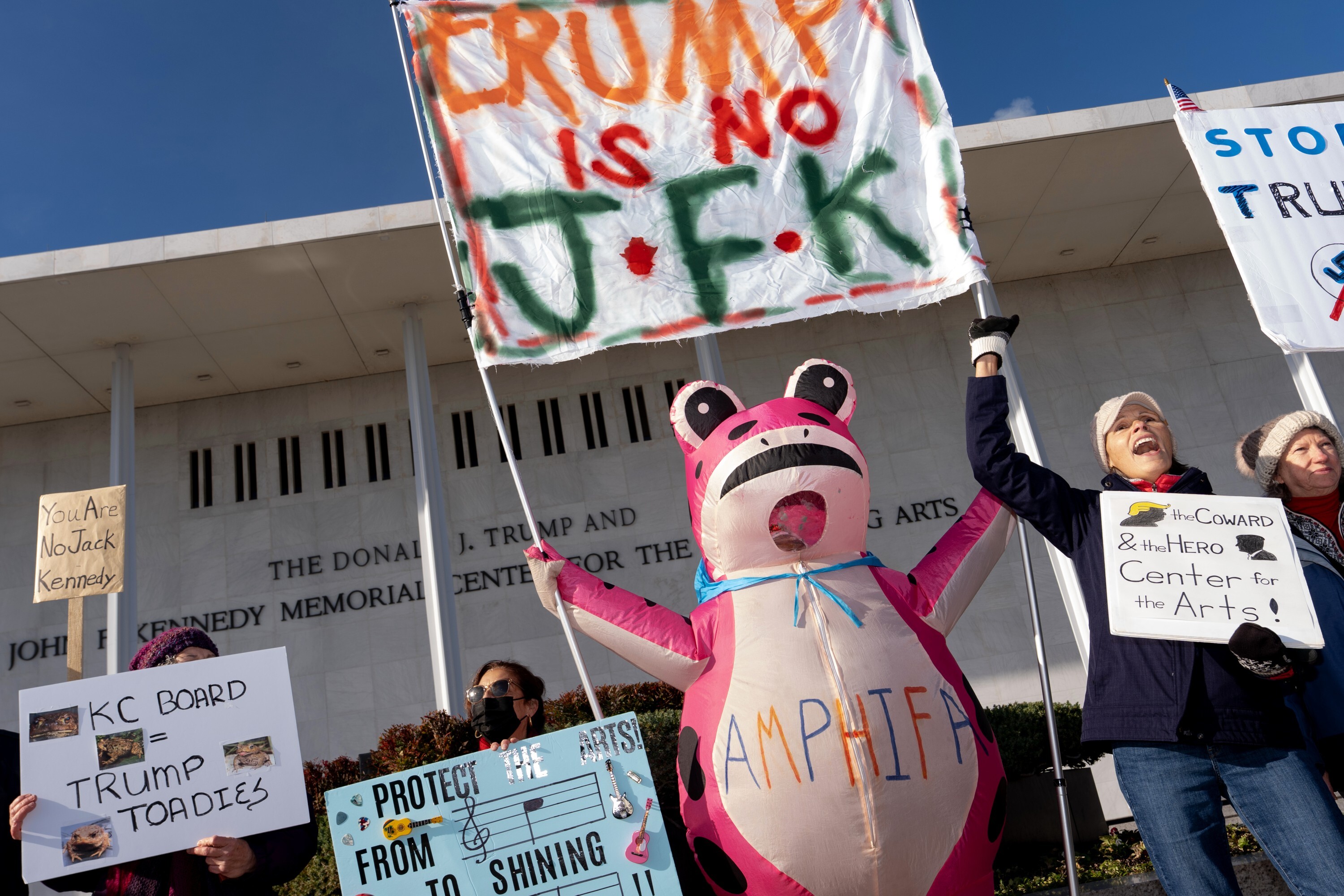 Demonstrators, including Nadine Siler, of Waldorf, Md., dressed in a pink frog costume, hold up signs at a designated protest point in front of the John F. Kennedy Memorial Center for the Performing Arts, a day after a Trump-appointed board voted to add President Donald Trump's name to the Kennedy Center, Saturday, Dec. 20, 2025, in Washington. (AP Photo/Julia Demaree Nikhinson)